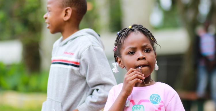 Girl biting her pendant with boy in grey hoodie behind her.