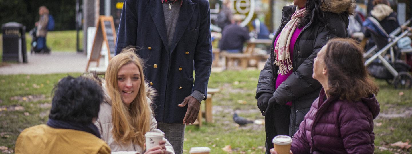 People gathered around an outdoor table drinking coffee and chatting, dressed in winter clothing.